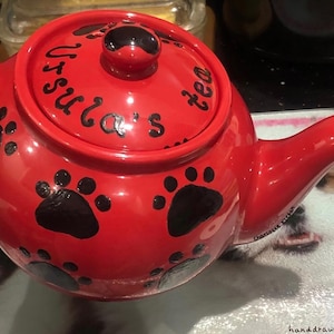 Könnte beinhalten: Rote Keramik-Teekanne mit schwarzen Pfotenabdrücken und dem Text "Ursula's tea" auf dem Deckel. Die Teekanne hat einen gebogenen Ausguss und einen Henkel. Ein Logo ist unten links zu sehen.