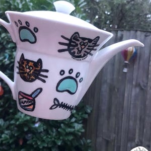 May include: A white ceramic teapot with a lid, decorated with hand-drawn illustrations of cat faces, light blue paw prints, a fish skeleton, and a can labelled Salmon. The text Charlotte Kleban is printed near one of the cat face illustrations.