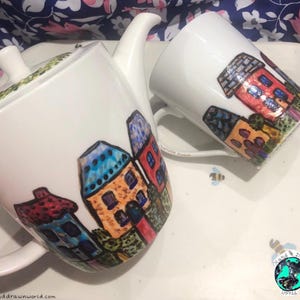 May include: A white teapot and mug with a hand-painted design of colourful houses. The teapot has a lid and a handle. The mug has a handle. Both pieces are decorated in a whimsical, cartoon-like style.