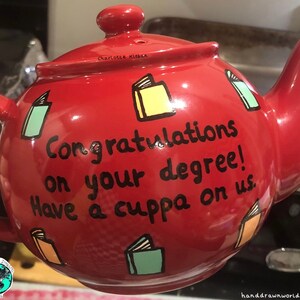 May include: Red teapot with a black inscription that reads "Congratulations on your degree! Have a cuppa on us." The teapot is decorated with colourful book illustrations.