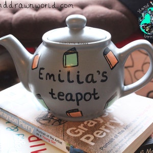 May include: A grey teapot with the words "Emilia's teapot" painted on it. The teapot is decorated with colourful book illustrations.