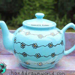 May include: A light blue ceramic teapot with a lid and handle. The teapot is decorated with a black and yellow floral pattern. The text "CHARLOTTE KLEBAN" is on the lid. The teapot is on a purple surface.