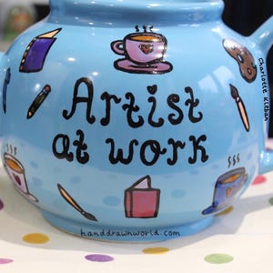 Könnte beinhalten: Eine hellblaue Teekanne mit handgemaltem Design, das den Text "Artist at Work" und verschiedene künstlerische Gegenstände wie Pinsel, Bleistifte, Bücher und Kaffeetassen zeigt. Die Teekanne ist mit einem schwarzen Umriss verziert und hat einen weißen Henkel.