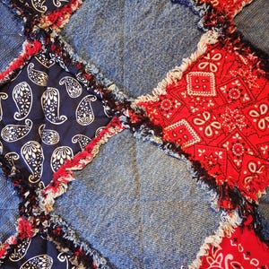 May include: A patchwork quilt featuring square panels of blue denim, navy blue fabric with a white paisley pattern, and red fabric with a white bandana print. The squares are joined with exposed, frayed seams that create a textured, grid-like appearance.