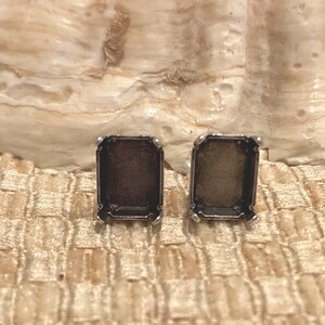 May include: Two silver earring settings with a rectangular shape and a beveled edge. The settings are empty and ready to be filled with stones or other embellishments.