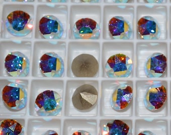 May include: A tray of iridescent, faceted rhinestones in various colors, including blue, red, and yellow. Two dull, gray, rounded stones are also present. The rhinestones are arranged in a grid pattern within a white plastic tray.