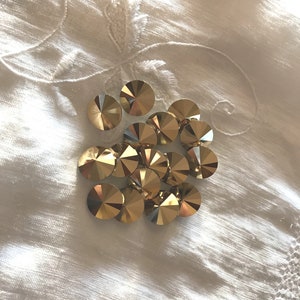 May include: A group of gold-colored, faceted, round rhinestones on a white fabric background.