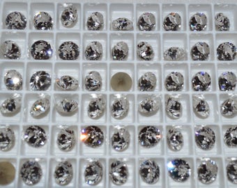 May include: A tray filled with numerous clear, faceted gemstones, each reflecting light. The stones vary in shape and size, with a few round and oval cuts. One stone is a different color. The tray is divided into small, square compartments.