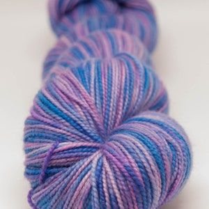 May include: Close-up of a skein of yarn in shades of blue, purple, and white. The yarn is tightly wound, with visible strands creating a textured appearance. The color transitions create a gradient effect, suitable for knitting or crochet projects.