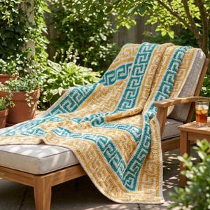 May include: A crocheted blanket with a geometric pattern in teal, yellow, and white is draped over a wooden lounge chair. The blanket's design features a repeating Greek key motif. The chair is outdoors, with greenery in the background.