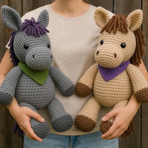 May include: Two handmade crochet stuffed animals: a gray donkey with purple hair and a green bandana, and a tan horse with brown hair and a purple bandana. Both are held in front of a wooden background.
