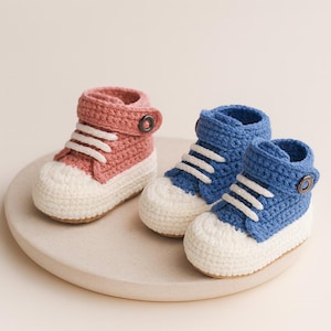 May include: Three crocheted baby sneakers in a studio setting. One pink sneaker and two blue sneakers are displayed. Each sneaker has white laces and a white toe cap. The pink sneaker has a brown button closure. The sneakers are on a round, light-colored surface.