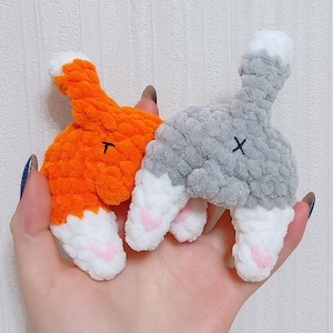 Op de afbeelding: Twee handgemaakte, pluche kattenkontornamenten. Eén is oranje met een zwarte "T" en witte en roze accenten. De andere is grijs met een zwarte "X" en witte en roze accenten. De ornamenten zijn gemaakt van een zacht, getextureerd materiaal.