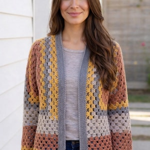 May include: A colorful, open-front crochet cardigan in shades of gray, yellow, and brown. The long-sleeved sweater features a geometric pattern and a gray border. The cardigan is worn over a gray t-shirt and blue jeans.