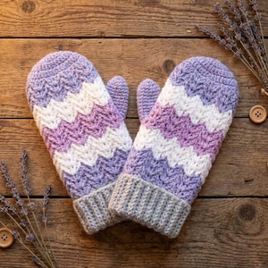 May include: A pair of handmade crochet mittens in a chevron pattern. The mittens feature alternating stripes of lavender, white, and light purple. The cuffs are a solid gray color. The mittens are displayed on a wooden surface.