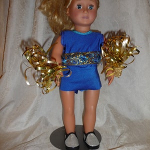 May include: A doll wearing a blue and gold cheerleading uniform with gold pom-poms. The doll has blonde hair and blue eyes.
