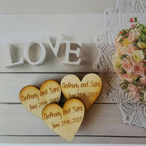 May include: Three wooden heart-shaped ornaments with the text "Anthony and Sara June 24th, 2017" engraved on each. The word "LOVE" is spelled out in white letters on a wooden surface.