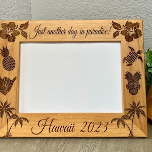 May include: A wooden picture frame with a white mat and a cutout design of tropical flowers, a pineapple, a hand gesture, a turtle, a tiki mask, and palm trees. The frame says "Just another day in paradise!" and "Hawaii 2023".
