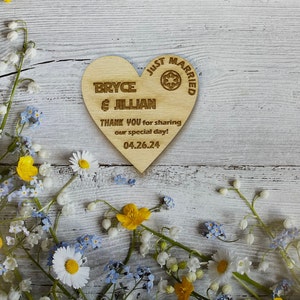 May include: A wooden heart-shaped sign with the text "Just Married" and a Star Wars symbol. The sign also includes the names "Bryce & Jillian" and the date "04.26.24". The sign is surrounded by white flowers.