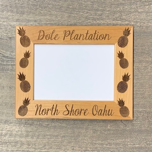 May include: A wooden picture frame with a light brown finish. The frame is decorated with pineapple designs and the text "Dole Plantation North Shore Oahu".