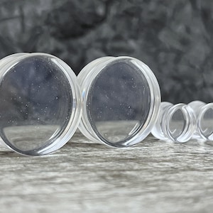 May include: Clear, transparent ear plugs in various sizes. The plugs are made of a smooth, polished material and have a rounded, circular shape. The image shows two large plugs and two smaller plugs.