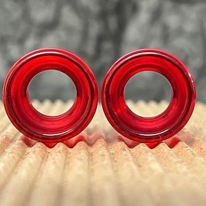 May include: A pair of circular ear plugs made from translucent red material. Each plug features a smooth, polished finish with a hollow center, creating a distinct ring shape. The vibrant red color is consistent across both pieces, which are displayed side by side.
