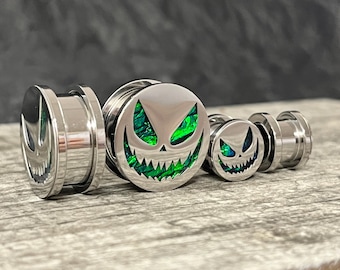 May include: A set of three silver-toned metal ear plugs featuring a circular face with a cut-out design of a grinning, sharp-toothed face. The cut-out sections are filled with a shimmering, iridescent green inlay that catches the light.