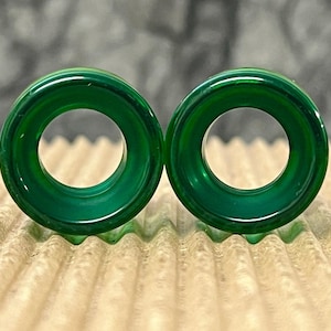 May include: A pair of circular ear plugs made from a smooth, translucent deep green material. Each plug features a wide, flat front face with a hollow center, showcasing a polished finish that reflects light across the rounded edges of the jewelry.