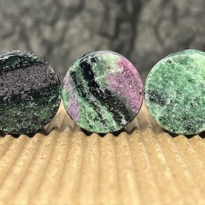 May include: Three circular ear plugs crafted from zoisite stone, each featuring a unique pattern of deep green, black, and magenta mineral inclusions. The stones have a polished, smooth surface and are arranged in a horizontal row.