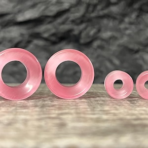 May include: Four pink, circular ear gauges of varying sizes are arranged on a wooden surface. The gauges have a smooth, polished appearance and a hole in the center. The color is a soft, translucent pink.