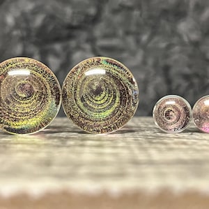 May include: A collection of four clear, round ear plugs with a galaxy-like design. The plugs vary in size, with two larger plugs and two smaller ones. The design features swirling patterns in shades of gold, purple, and green.