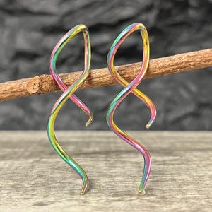 May include: A pair of iridescent spiral earrings. The earrings are a mix of colors, including green, yellow, pink, and purple. They are curved and have a smooth, metallic finish. The earrings are displayed on a wooden branch.