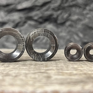 May include: A collection of dark gray and silver-toned ear gauges. The set includes two large tunnel-style gauges and two smaller plugs. The gauges have a polished, reflective surface with a subtle, linear pattern. The jewelry is displayed on a light-colored, textured surface.
