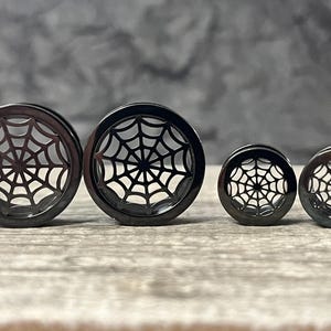 May include: Four black circular ear plugs arranged in a row, featuring a cut-out spider web design. The two larger plugs are on the left, and the two smaller plugs are on the right. Each piece has a smooth, polished finish and a hollow center.