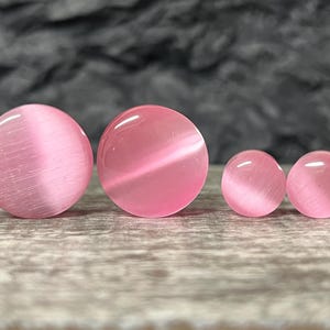May include: Four pink cat's eye stone plugs of varying sizes. The plugs are round with a polished finish and a visible band of light across the center. The background is a textured gray surface.