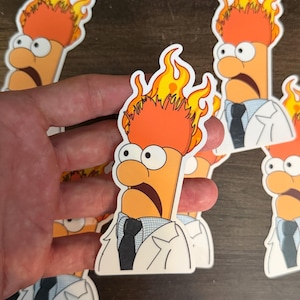 May include: A die-cut vinyl sticker featuring a cartoon character with orange skin, wide eyes, and a mouth open in shock. The character has bright orange and yellow flames erupting from their head and wears a white lab coat over a blue shirt and tie.