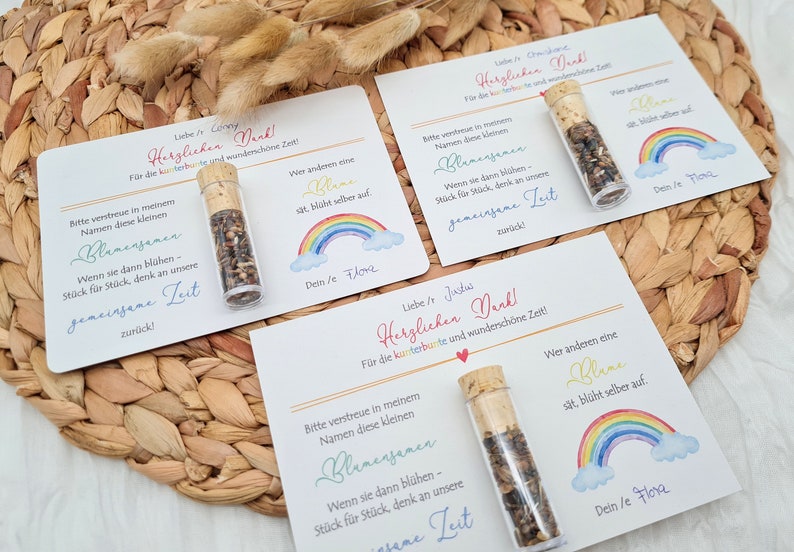 K&ouml;nnte beinhalten: Drei kleine Glasfl&auml;schchen mit Korkstopfen, gef&uuml;llt mit Blumensamen. Jedes Fl&auml;schchen ist an einer wei&szlig;en Karte mit einem Regenbogenmuster und dem Text "Herzlichen Dank!"  "Bitte verstreue in meinem Namen diese kleinen Blumen Samen - Wenn sie dann bl&uuml;hen - St&uuml;ck f&uuml;r St&uuml;ck, denk an unsere gemeinsame Zeit zur&uuml;ck!" und "Wer anderen eine Blume s&auml;t, bl&uuml;ht selber auf. Dein/e Flora" befestigt.
