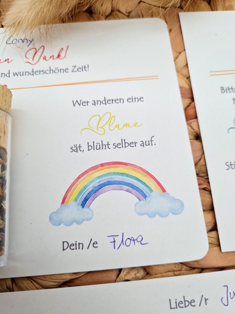 K&ouml;nnte beinhalten: Eine wei&szlig;e Karte mit einem Regenbogen, Wolken und dem Text "Wer anderen eine Blume s&auml;t, bl&uuml;ht selber auf." Die Karte enth&auml;lt auch den Text "Dein/e Flora" und "Liebe/r Ju".