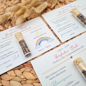 K&ouml;nnte beinhalten: Zwei wei&szlig;e Karten mit buntem Text und einer Regenbogen-Illustration. Der Text auf den Karten lautet "Herzlichen Dank!" was im Deutschen "Vielen Dank!" bedeutet. Jede Karte hat ein kleines Glasfl&auml;schchen mit Blumensamen. Die Fl&auml;schchen haben Korkstopfen.