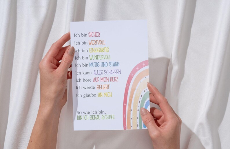 May include: A white card with a rainbow design and the text "Ich bin SICHER, Ich bin WERTVOLL, ich bin EINZIGARTIG, Ich bin WUNDERVOLL, Ich bin MUTIG UND STARK, Ich kann ALLES SCHAFFEN, Ich h&ouml;re AUF MEIN HERZ, Ich werde GELIEBT, Ich glaube AN MICH, So wie ich bin, BIN ICH GENAU RICHTIGI" in German.