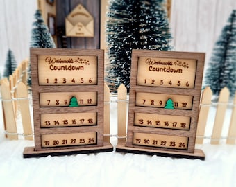 May include: Two wooden Christmas countdown calendars with the text "Weihnachts Countdown" and numbers 1-24. Each calendar has a small green Christmas tree icon. The calendars are set against a backdrop of miniature Christmas trees and a white picket fence.