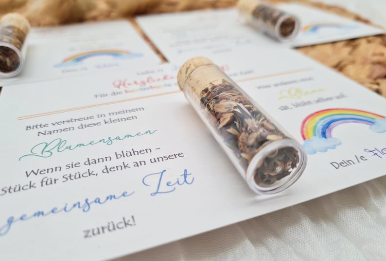 K&ouml;nnte beinhalten: Eine wei&szlig;e Karte mit einem Regenbogen und Wolken darauf gedruckt. Die Karte enth&auml;lt den Text "Bitte verstreue in meinem Namen diese kleinen Blumensamen. Wenn sie dann bl&uuml;hen - St&uuml;ck f&uuml;r St&uuml;ck, denk an unsere gemeinsame Zeit zur&uuml;ck!" und "Dein/e Flo". Ein kleines Glasr&ouml;hrchen gef&uuml;llt mit Blumensamen liegt auf der Karte.