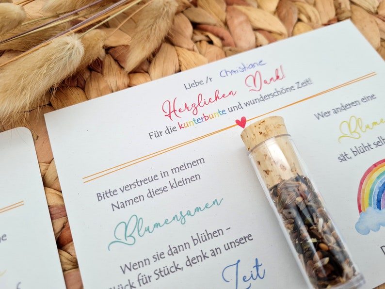 K&ouml;nnte beinhalten: Eine wei&szlig;e Karte mit einer handschriftlichen Nachricht auf Deutsch, die sich f&uuml;r die Zeit bedankt. Die Karte ist mit einem kleinen Glasfl&auml;schchen gef&uuml;llt mit Blumensamen, einem Regenbogen und einem Herzen verziert.