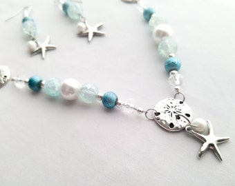 May include: A silver necklace with blue and white beads, a silver starfish charm, and a silver sand dollar charm. The necklace is on a white background.