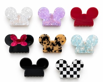 May include: A collection of seven hair clips in the shape of a mouse head. The clips come in various colors and patterns, including white, purple, red, black with a pink bow, tortoiseshell, blue, and black and white checkered.