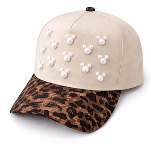 May include: A beige baseball cap with a leopard print brim. The cap is adorned with multiple pearl-like embellishments arranged in a Mickey Mouse head shape. The cap has a brown button on top.