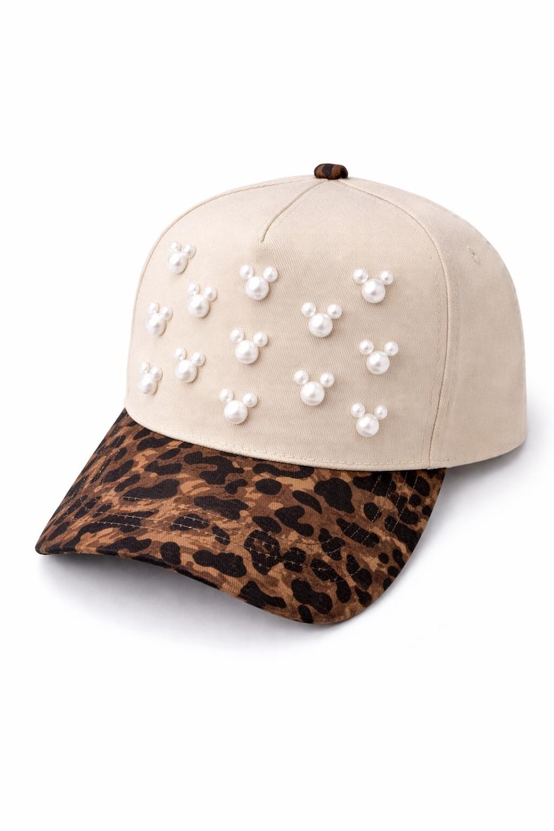 May include: A beige baseball cap with a leopard print brim. The cap is adorned with multiple pearl-like embellishments arranged in a Mickey Mouse head shape. The cap has a brown button on top.