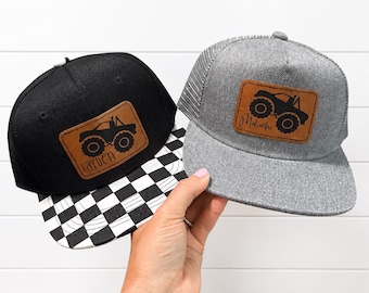 May include: A black baseball cap with a checkered brim and a brown leather patch featuring a monster truck and the name Cayden, alongside a grey trucker hat with a brown leather patch featuring a monster truck and the name Malachi.