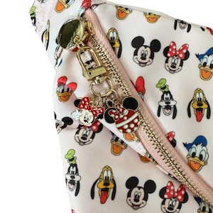 May include: A light pink cosmetic bag with a gold zipper and charm, featuring a pattern of Mickey Mouse, Minnie Mouse, Donald Duck, and Goofy. The bag has the text "Add Bag Charm!" in black.