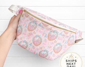 May include: A pink fanny pack featuring a repeating pattern of ornate gold-framed castle illustrations, blue bows, and small teacups. The bag has a gold-tone metal zipper closure and an adjustable white strap. A circular badge displays the text SHIPS NEXT DAY.